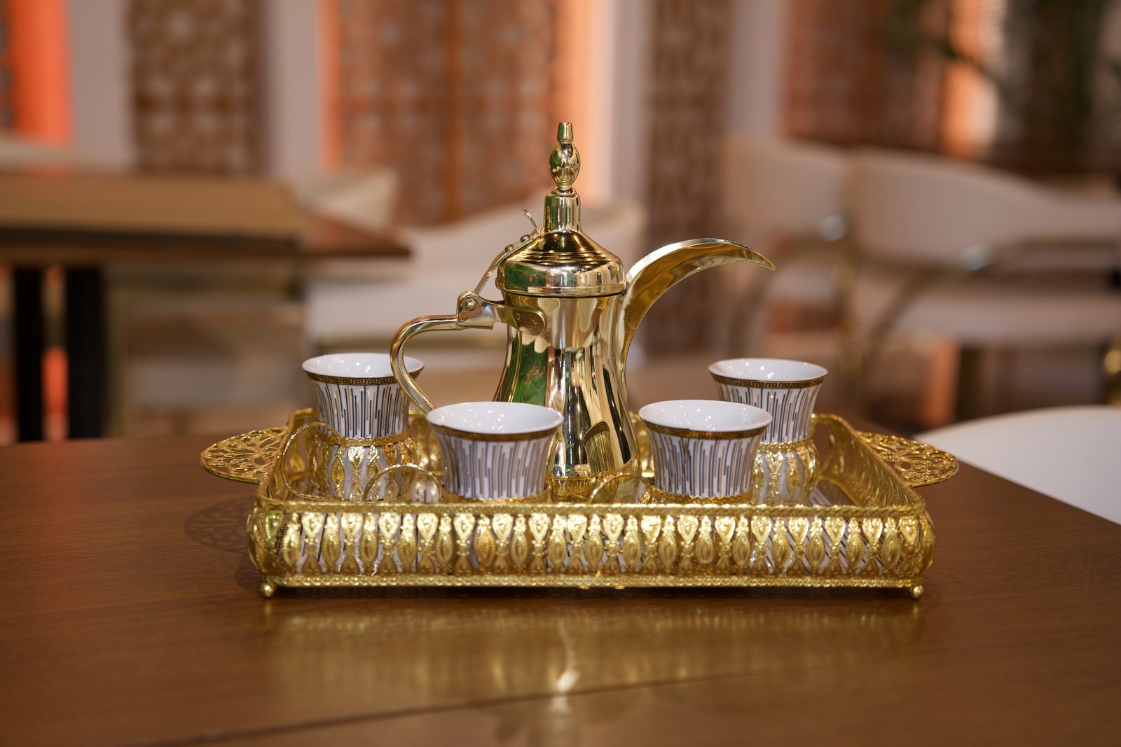 A gold dallah coffee pot and handleless cups on an ornate tray before mashrabiya arches