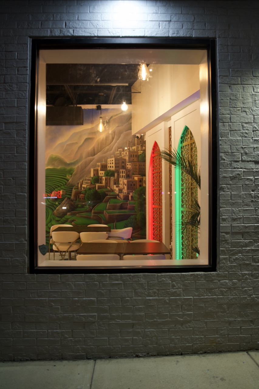 Qahwah Drip at night — the green mountain-village mural and colorfully lit arches glimpsed through the window